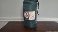 Green Woods sleeping bag in storage sack on wooden table, front view showing product label