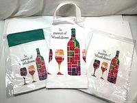 Three canvas wine gift bags displayed, two with green fold-over tops inside plastic sleeves, and one with printed cotton kitchen towel in the center showing colorful abstract wine bottle and glass design with text.
