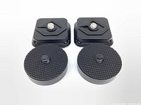 Two black square and round quick release plates, viewed from top showing textured mounting surfaces and screws.