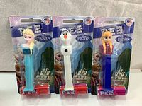 Three PEZ dispensers in original sealed packaging featuring Elsa, Olaf, and Anna from Disney Frozen with candy attached.
