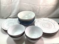 Photo shows two stacks of white plates with embossed patterns, two white bowls with brown rims, and a group of multi-colored large and medium bowls.