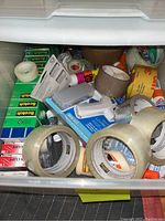 Drawer with various adhesive tapes including clear packing, brown packing tape rolls, Scotch tape, glue stick, and other adhesives.