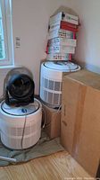 Two white Honeywell air purifiers stacked with a small black fan on top and a cardboard box beside them. Stacked boxes of HEPA filters atop one purifier.
