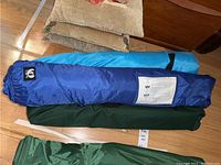 Photo showing three packed bags on a hardwood floor. Bags are green and two shades of blue, indicating different items inside. A partially visible label is on the floor under the green bag.