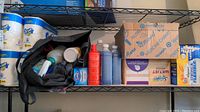 Shot of metal shelf with cleaning products, paper towels, and boxed items including facial tissues and trash bags, plus various bottles in a black bag
