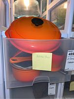 Set of Le Creuset orange enamel cookware in plastic bin showing pots and pan stacked inside, with lids and handles visible.