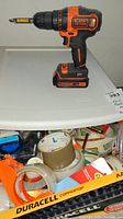 Orange and black Black & Decker cordless drill with lithium-ion battery on top of bin with many rolls of tape and some battery packs.