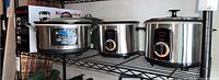 Kitchen appliances and cookware on metal shelf: Hamilton Beach slow cooker, Pars rice cookers, double steam pot with lid.