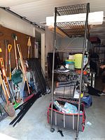 Three metal wire shelving units visible. One fully assembled with items on it and wheels attached. Background shows garage tools and other items.