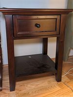 Front view of one wooden nightstand showing single drawer and lower shelf with dust on the surface.