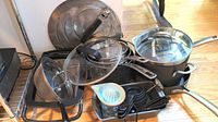 Overview image showing various pots, pans, glass lids, measuring cups, spoons, juicer, and strainer arranged on floor.