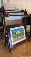 Wooden quilt rack and signed framed art placed in front of it on a wooden floor.
