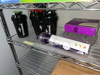 Shelf with four Kitchenie brand black shaker bottles and a purple plastic measuring tool.