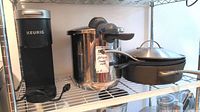 Photo showing Keurig coffee maker, WMF Perfect Plus pressure cooker, Calphalon pans, and Lodge cast iron pan on metal shelving