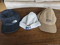 Photo shows three hats placed on a wooden surface: black insulated cap (The North Face), cream knit beanie (The North Face), and brown Patagonia cap.