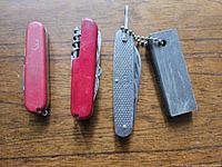 Three vintage pocket cutters displayed on wood surface: red Swiss Army knife with multiple tools, silver US Army pocket knife, and a rectangular metal sharpening stone on a chain.