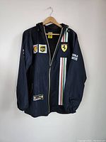 Front view of black Ferrari team waterproof jacket with chest sponsor logos and Italian flag striping, hanging on wooden hanger.