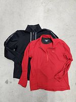 Two men's tops laid flat on a concrete surface: a red Arcteryx pullover and a black Lululemon long sleeve top.