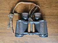 Front view of black textured vintage German binoculars with attached worn leather strap.