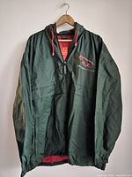 Front view of dark green Jaguar Racing windbreaker jacket hanging on wooden hanger, showing red drawstrings, half-zip, and front pocket.