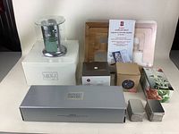 Photo showing assortment of kitchenware including Mikasa candle, cookbook stand, cutting boards, Royal Doulton ladle, mesh containers, floral dish, and boxed items on white surface.