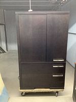 Frontal view of the espresso coloured wardrobe on a dolly, showing two vertical doors at the top and three drawers below with metal handles.