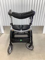 Front view of folding walker showing padded handlebar, dual padded seat, large front wheels, and mesh storage basket beneath seat