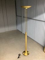 Full view of gold floor lamp showing round base, slender vertical structure, conical lampshade and attached power cord.