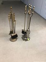 Two sets of metal fireplace tools placed side by side on a concrete surface. Each set includes a stand and four tools: poker, shovel, tongs, and brush with long handles and rounded tops.