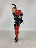 Front and side views of Mephistopheles porcelain figurine standing on a white round base, showing painted red and black clothing, facial detail, and chipped hand.