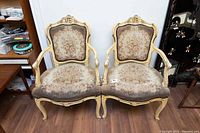 Front view of two antique arm chairs side by side showing carved wood and floral upholstery with wear.