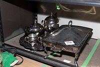 Photo showing entire vintage silver plated 5-piece tea set arranged on a serving tray next to the footed covered chafing dish with lid closed on a metal shelf.