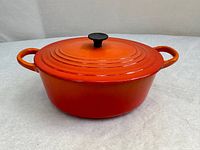 Front and side view of vibrant orange Le Creuset oval Dutch oven with lid, showing loop handles and knob on lid.