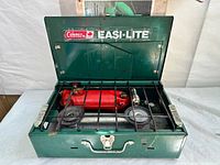 Open Coleman Easi-Lite camp stove showing two burners with black grate and red fuel tank inside.
