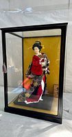 Full view of the Geisha doll inside the wood and glass display case from the front.