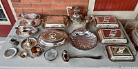 Overall front view of the full assortment of silver plated serveware laid out on table against brick background