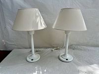 Pair of white table lamps shown side by side, both plugged in with cords, on a white surface against a white backdrop.