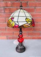 Full view of stained glass style table lamp showing cream and floral patterned shade with red glass accent on metal base.