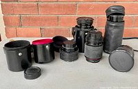 Five vintage camera lenses and multiple black protective cases with red velvet lining, photographed on a flat surface against a brick backdrop.