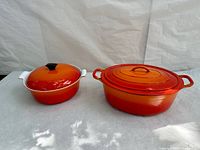 Two vintage orange enamel cast iron cookware pieces with lids, one oval and one round, shown together.