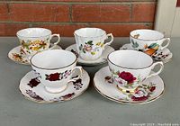 Front view of 5 bone china cups and saucers arranged in two rows showing detailed floral patterns and gold trims.