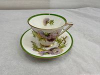 Fine bone china tea cup and saucer set with purple thistle floral design and green and gold rims, shown together.