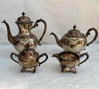 Full view of the four-piece silver plated tea and coffee set showing coffee pot, teapot, sugar bowl with lid, and creamer, all with ornate design and patina.