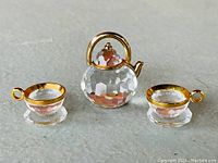 Front view of clear faceted crystal teapot with gold-tone handle and two matching crystal teacups with saucers