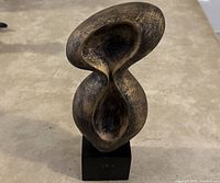 Front view of abstract sculpture art piece with concave flowing organic shapes on a black pedestal placed on a beige carpet.