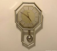 Octagonal wall clock with aluminum frame and glass front, featuring a lower glass panel with a silver pendulum. Clock face includes the Cardinal brand name and simple hour markers.