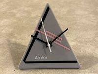 Photo of a triangular contemporary grey clock made of hard plastic with black and pink geometric design lines and three clock hands, no numbers visible.