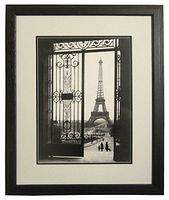 Framed black and white photographic print depicting the Eiffel Tower seen through ornate iron gates, matted white and enclosed in a black wooden frame with glass, wire hanger on back.