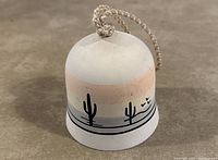 White hanging bell with painted Saguaro cactus silhouettes and birds on a pastel stripe background, showing braided hanging cord.