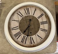 Full view of large round clock with vintage style face, Roman numerals, and white metallic frame on table surface